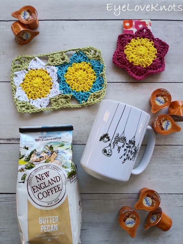 Lily's blue and white Floral Mug Rug paired together with Lily's pink Floral Gift Card Holder, a bag of New England Coffee Butter Pecan Flavor, a Hanging Plants coffee mug, and Hazelnut coffee creamers, EyeLoveKnots