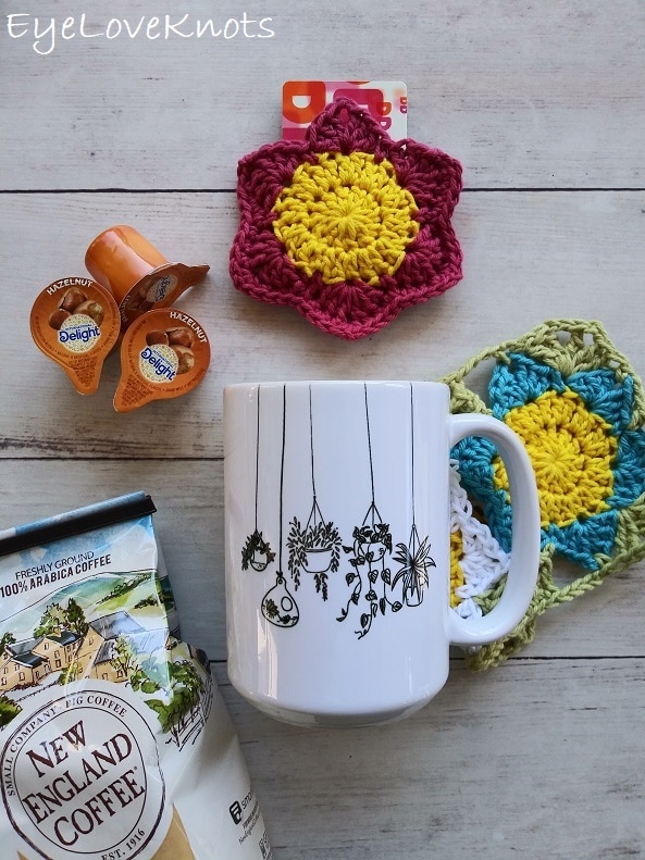 Lily's blue and white Floral Mug Rug paired together with Lily's pink Floral Gift Card Holder, a bag of New England Coffee Butter Pecan Flavor, a Hanging Plants coffee mug, and Hazelnut coffee creamers, EyeLoveKnots